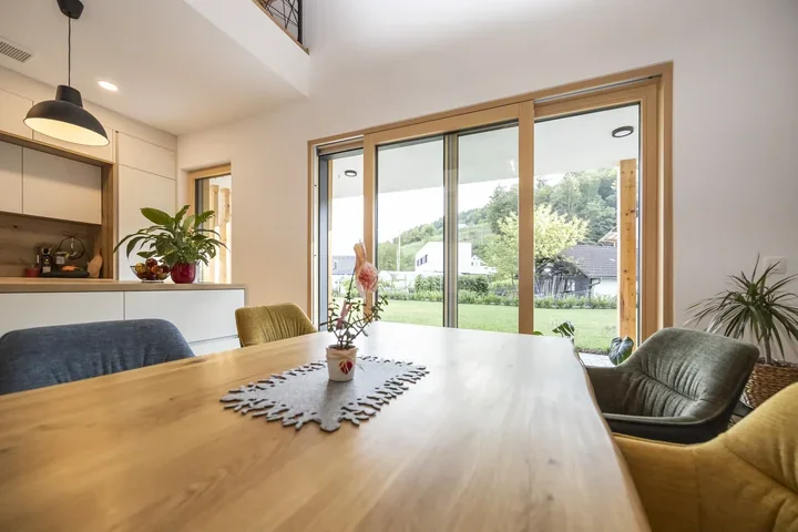 Jedilnica v montažni hiši v Kamnici z lesenimi okvirji velike zasteklitve in pogledom na vrt.