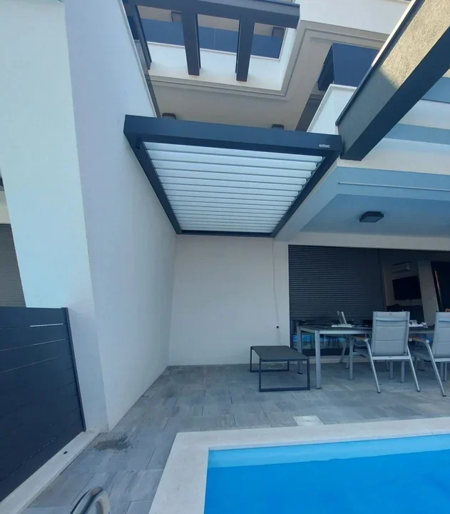 Temna pergola s svetlimi lamelami ob beli fasadi nad zunanjo jedilno mizo in bazenom.